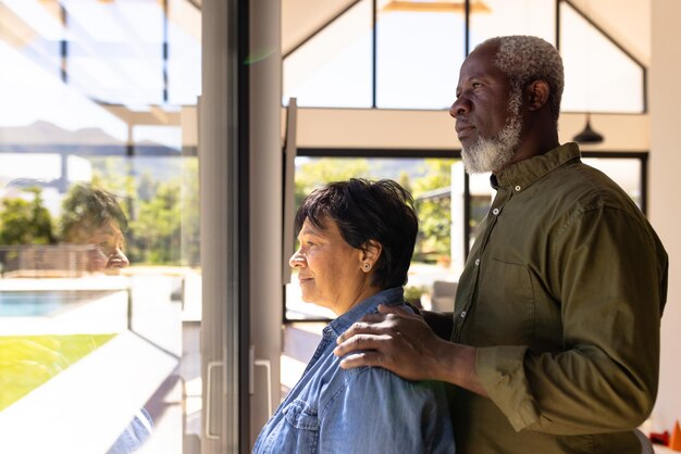 Foto vista lateral de um casal sênior multirracial olhando pela janela em pé na casa de repouso. sério, inalterado, amor, união, apoio, vida assistida e conceito de aposentadoria.