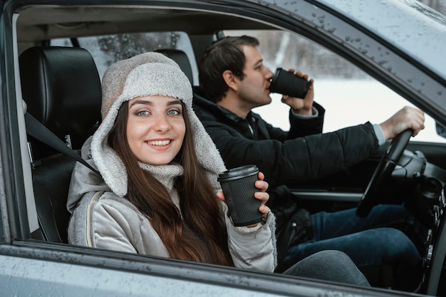 Foto vista lateral de um casal sorridente no carro tomando uma bebida quente durante uma viagem