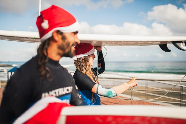 Foto vista lateral de um casal vestindo chapéus de papai noel carregando pranchas de surf nas cabeças