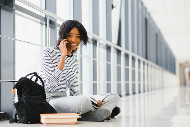 Vista lateral de um estudante afro-americano sentado no chão e lendo um livro no corredor