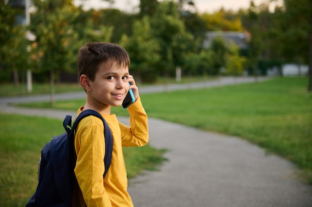 Vista lateral de um estudante vestindo um moletom amarelo com mochila andando no caminho em um parque público, indo para casa depois da escola, falando no celular, sorrindo com um sorriso cheio de dentes para a câmera