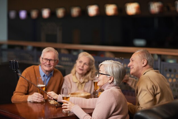 Vista lateral de um grupo de idosos modernos tirando uma foto de selfie enquanto bebe cerveja no bar e aproveita a noite com os amigos, copie o espaço