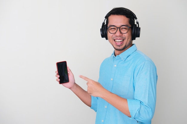 Vista lateral de um homem adulto asiático usando fone de ouvido, mostrando uma expressão feliz e apontando para o seu telefone celular