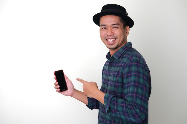 Vista lateral de um homem asiático descolado sorrindo feliz enquanto aponta para o seu telefone celular