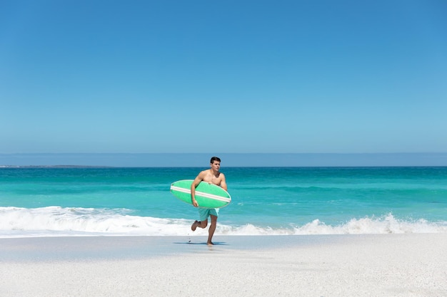 Vista lateral de um homem caucasiano correndo na praia com céu azul e mar ao fundo, carregando uma prancha de surf
