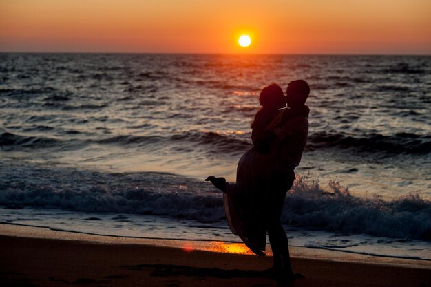 Foto vista lateral de um homem de silhueta beijando uma mulher na praia contra o céu durante o pôr do sol