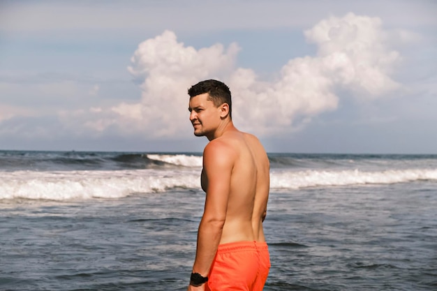 Vista lateral de um homem desportivo em shorts de pé no oceano contra o pano de fundo das ondas em um dia ensolarado