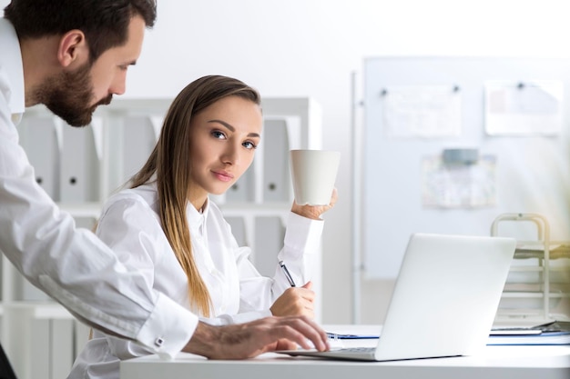 Vista lateral de um homem e uma mulher em um escritório branco. Eles estão trabalhando em um laptop. Ele está pressionando uma tecla. Ela está olhando para o espectador e segurando uma xícara de café