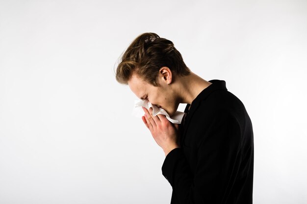 Foto vista lateral de um homem segurando um lenço de papel enquanto espirra contra um fundo branco