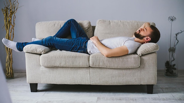 Vista lateral de um jovem barbudo bonito em roupas casuais dorme ou relaxa no sofá em uma sala em casa