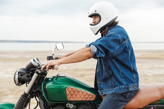 Vista lateral de um jovem bonito vestindo roupa casual, sentado em uma motocicleta na praia, usando um capacete