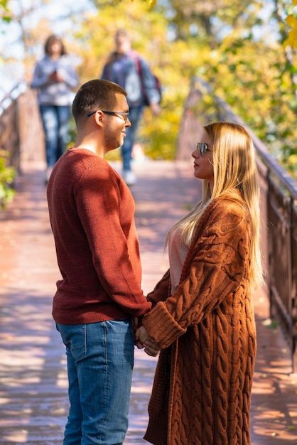 Vista lateral de um jovem casal moderno apaixonado, de mãos dadas e olhando um para o outro em uma ponte no parque