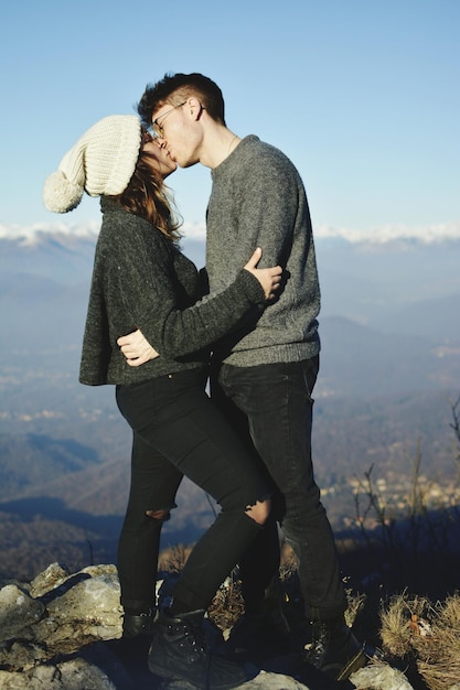Vista lateral de um jovem casal se beijando enquanto está de pé na montanha contra o céu