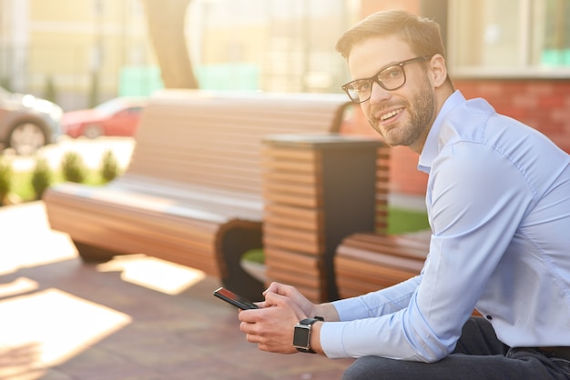 Vista lateral de um jovem empresário feliz segurando um smartphone, olhando para a câmera e sorrindo enquanto