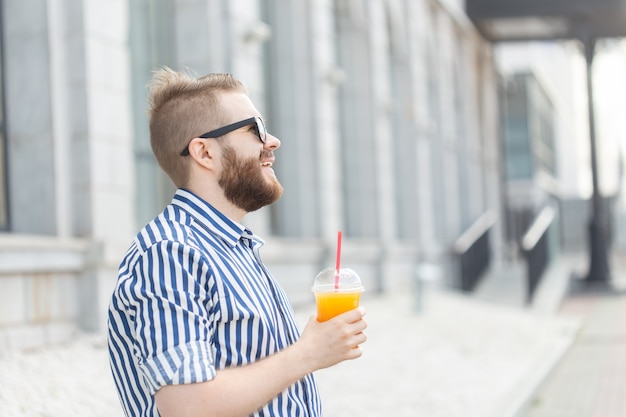 Vista lateral de um jovem empresário muito alegre, com um bigode elegante e uma barba com suco nas mãos, andando pela cidade após um dia de trabalho. Conceito de positivo e descanso.