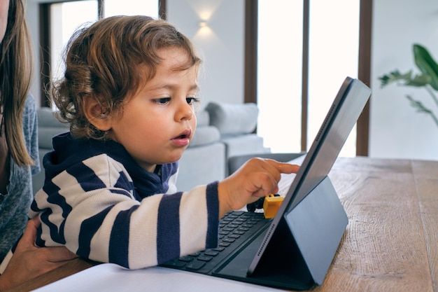 Vista lateral de um menino bonito sentado em uma mesa de madeira e navegando no laptop com a mãe da colheita em uma sala aconchegante