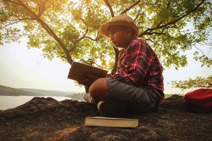 Foto vista lateral de um menino com um livro sentado no parque ao lado do lago