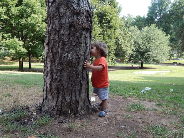 Vista lateral de um menino de pé junto ao tronco de uma árvore no parque