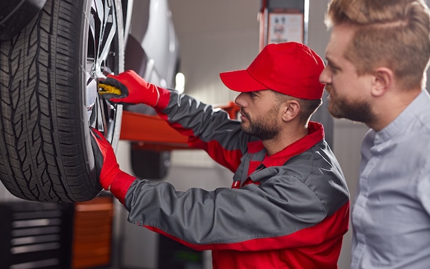 Vista lateral de um técnico de manutenção profissional com uma chave inglesa examinando a roda de um automóvel quebrado de um cliente do sexo masculino na estação de serviço