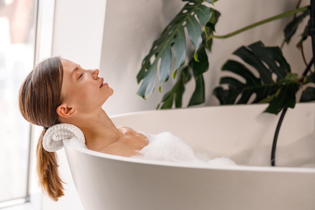 Vista lateral de uma bela jovem relaxando com os olhos fechados enquanto toma um banho de espuma no luxo