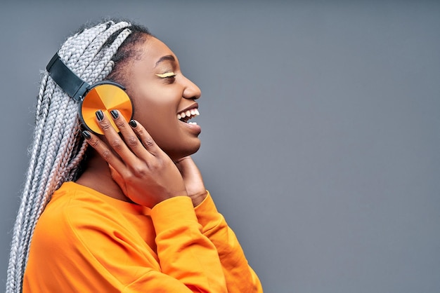 Vista lateral de uma jovem africana alegre ouvindo música com fones de ouvido isolados na parede cinza
