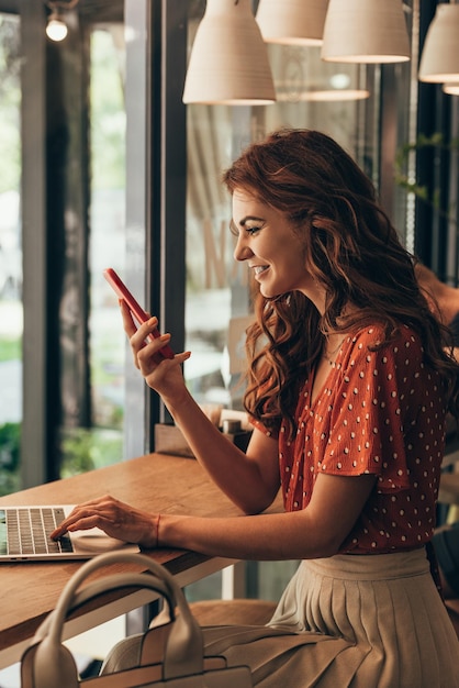 Foto vista lateral de uma jovem alegre usando um smartphone na mesa com um laptop na cafeteria