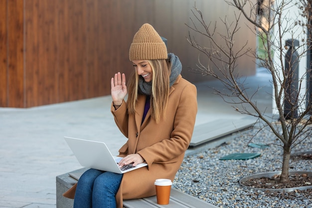 Vista lateral de uma jovem conversando on-line em um laptop sentado no banco, uma jovem freelancer conversando nas mídias sociais
