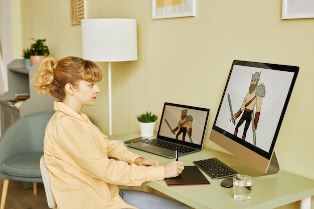 Vista lateral de uma jovem designer loira criando novas imagens digitais enquanto está sentada em frente ao des
