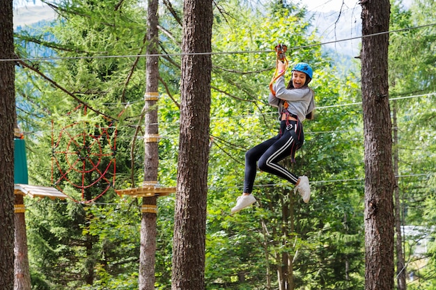 Vista lateral de uma jovem excitada com a boca aberta olhando para baixo enquanto se diverte em cordas altas com capacete protetor de cabos de segurança no parque de aventura florestal à luz do dia