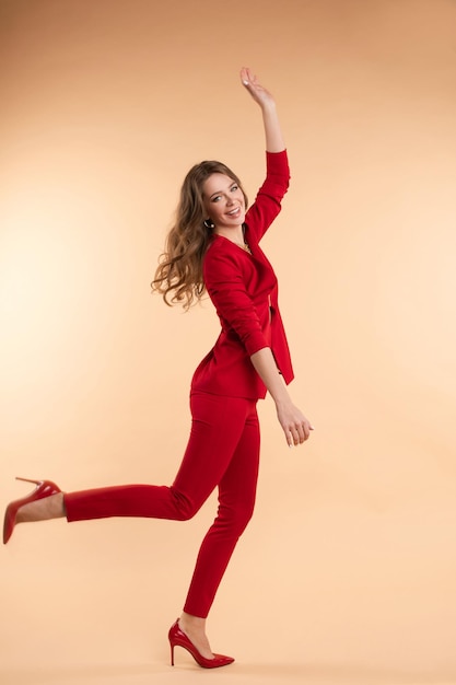 Vista lateral de uma jovem feliz de terno vermelho inteligente, olhando para a câmera e pulando no estúdio beautidul senhora de salto alto rindo e posando em fundo isolado conceito de beleza e felicidade