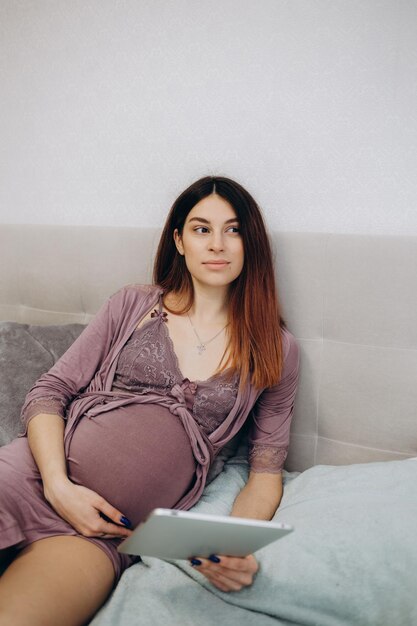 Vista lateral de uma jovem grávida descansando com um tablet no sofá em casa Uma linda mulher grávida que segura a barriga e relaxa com um tablet digital na cama durante a gravidez