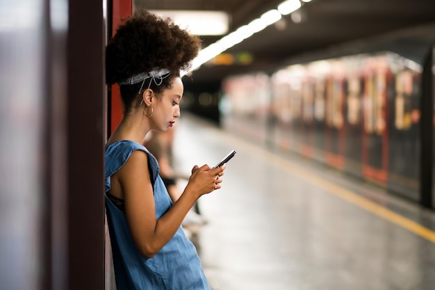 Vista lateral de uma jovem marroquina com faixa de cabelo afro olhando para a tela do celular enquanto estava verificando mensagens de texto na plataforma do metrô à noite e esperando o trem