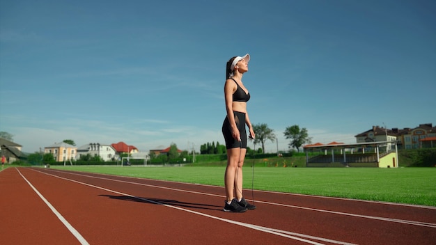 Vista lateral de uma jovem muscular cansada usando boné branco e roupa esportiva preta, descansando durante os exercícios de aquecimento no estádio. Menina apta com os olhos fechados, segurando a corda de pular ao ar livre.