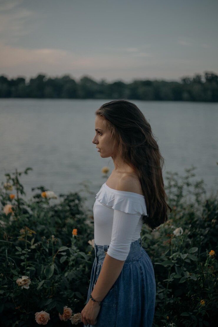 Foto vista lateral de uma jovem pensativa de pé junto a plantas contra o lago
