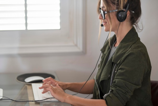 Vista lateral de uma jovem trabalhando no computador e usando fones de ouvido Fundo de estilo de vida de negócios de escritório Mulheres modernas escrevendo no teclado e sorrindo Trabalho de negócios on-line Trabalho de tecnologia