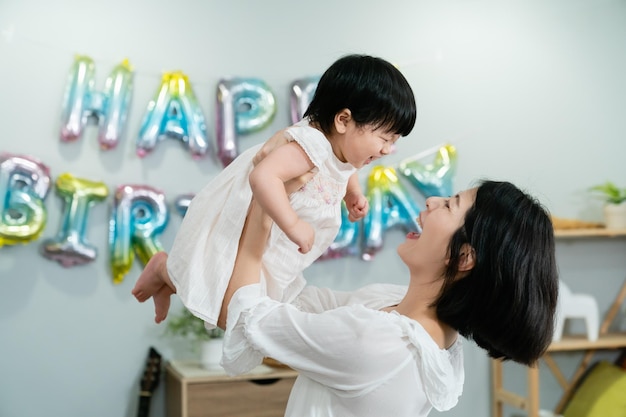 vista lateral de uma mãe coreana alegre segurando sua adorável filha bebê no alto com a parede decorada para aniversário no fundo.