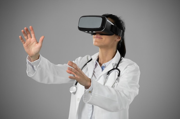 Vista lateral de uma médica caucasiana vestindo um jaleco com estetoscópio, usando um fone de ouvido de realidade virtual com as mãos tocando uma tela interativa virtual em um fundo cinza.