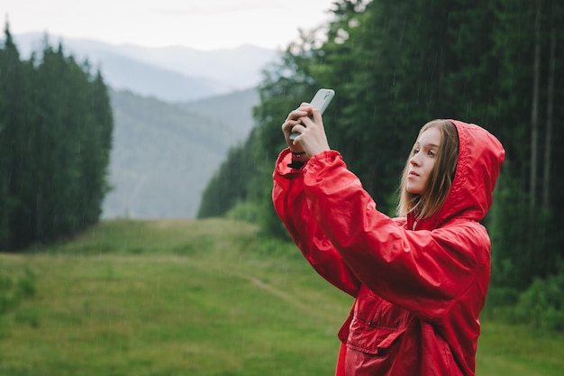 Vista lateral de uma mulher alpinista tomando selfie em seu celular usando uma capa de chuva vermelha