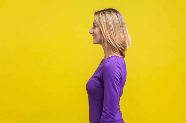 Vista lateral de uma mulher bem sucedida positiva no vestido roxo em pé com sorriso cheio de dentes, parecendo feliz e conteúdo vazio espaço de cópia para anúncio estúdio interno tiro isolado em fundo amarelo