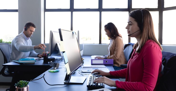 Foto vista lateral de uma mulher caucasiana trabalhando como suporte ao cliente, vestindo roupas elegantes e fones de ouvido, usando um computador desktop com colegas trabalhando em segundo plano.