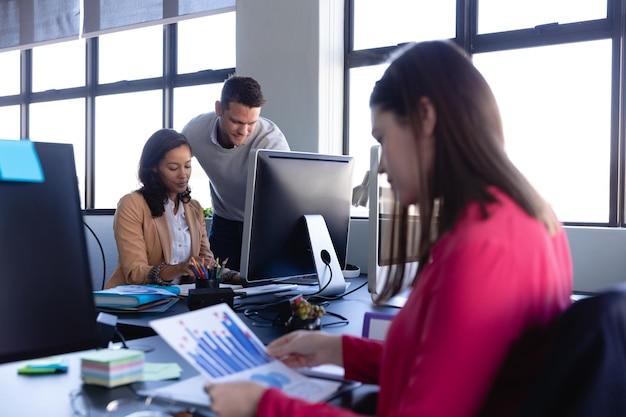 Vista lateral de uma mulher caucasiana trabalhando em um escritório moderno, olhando para um gráfico, com dois colegas trabalhando em segundo plano.