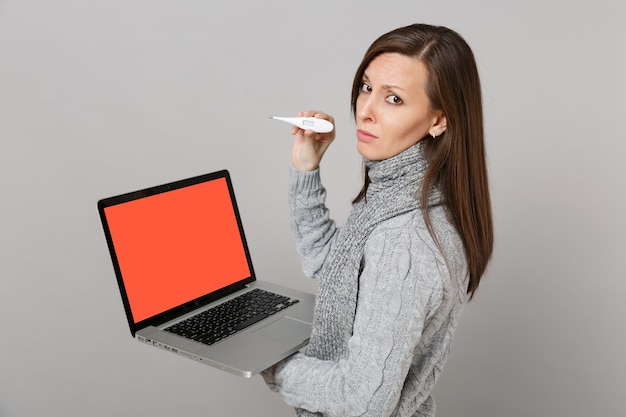 Vista lateral de uma mulher chateada em um cachecol de suéter segurando o termômetro laptop pc com tela vazia em branco isolada no fundo cinza. Tratamento on-line de estilo de vida saudável, consultando o conceito de estação fria.