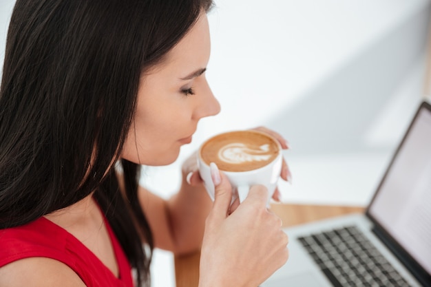 Vista lateral de uma mulher de camisa vermelha tomando café com os olhos fechados no local de trabalho