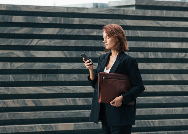 Vista lateral de uma mulher de negócios de meia-idade com uma pasta de couro e um smartphone caminhando ao ar livre