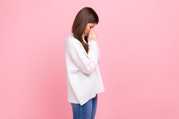 Vista lateral de uma mulher deprimida chorando, sentindo tristeza, estando preocupada e chateada, enxugue as lágrimas, vestindo um suéter branco estilo casual. tiro de estúdio interior isolado no fundo rosa.