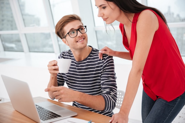 Vista lateral de uma mulher em pé perto de um homem sentado à mesa com o laptop no escritório