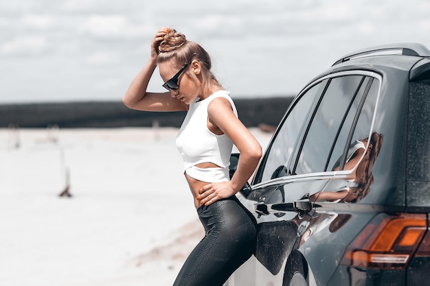Vista lateral de uma mulher graciosa magra em calças pretas e óculos de sol perto de carro preto olhando ao longo