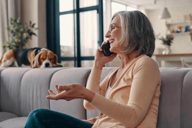 Vista lateral de uma mulher idosa bonita em roupas casuais, falando ao telefone inteligente e sorrindo enquanto está sentada no sofá em casa