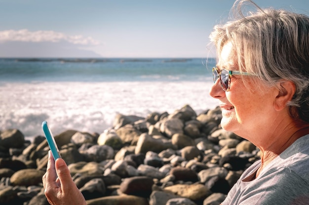 Vista lateral de uma mulher idosa sorridente usando telefone celular enquanto está sentada na praia ao pôr do sol idosa feliz desfrutando de tempo livre e aposentadoria no mar