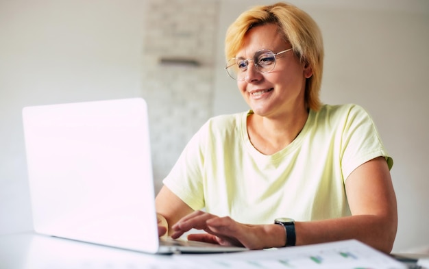 Vista lateral de uma mulher madura inteligente, confiante e feliz usando óculos de proteção usando um laptop enquanto trabalha remotamente de casa na sala de estar ou faz alguma compra on-line de casa